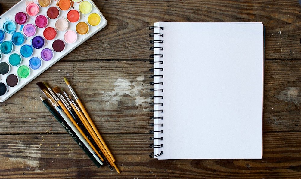 A sketch pad and some paints on a wooden table.